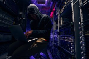 A person in a hooded jacket typing on a laptop in a darkened computer room.